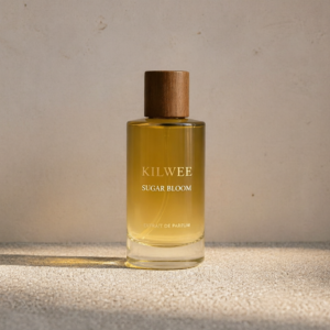 Perfume bottle with a wooden cap labeled KILWEE SUGAR BLOOM on a textured beige surface under warm light.